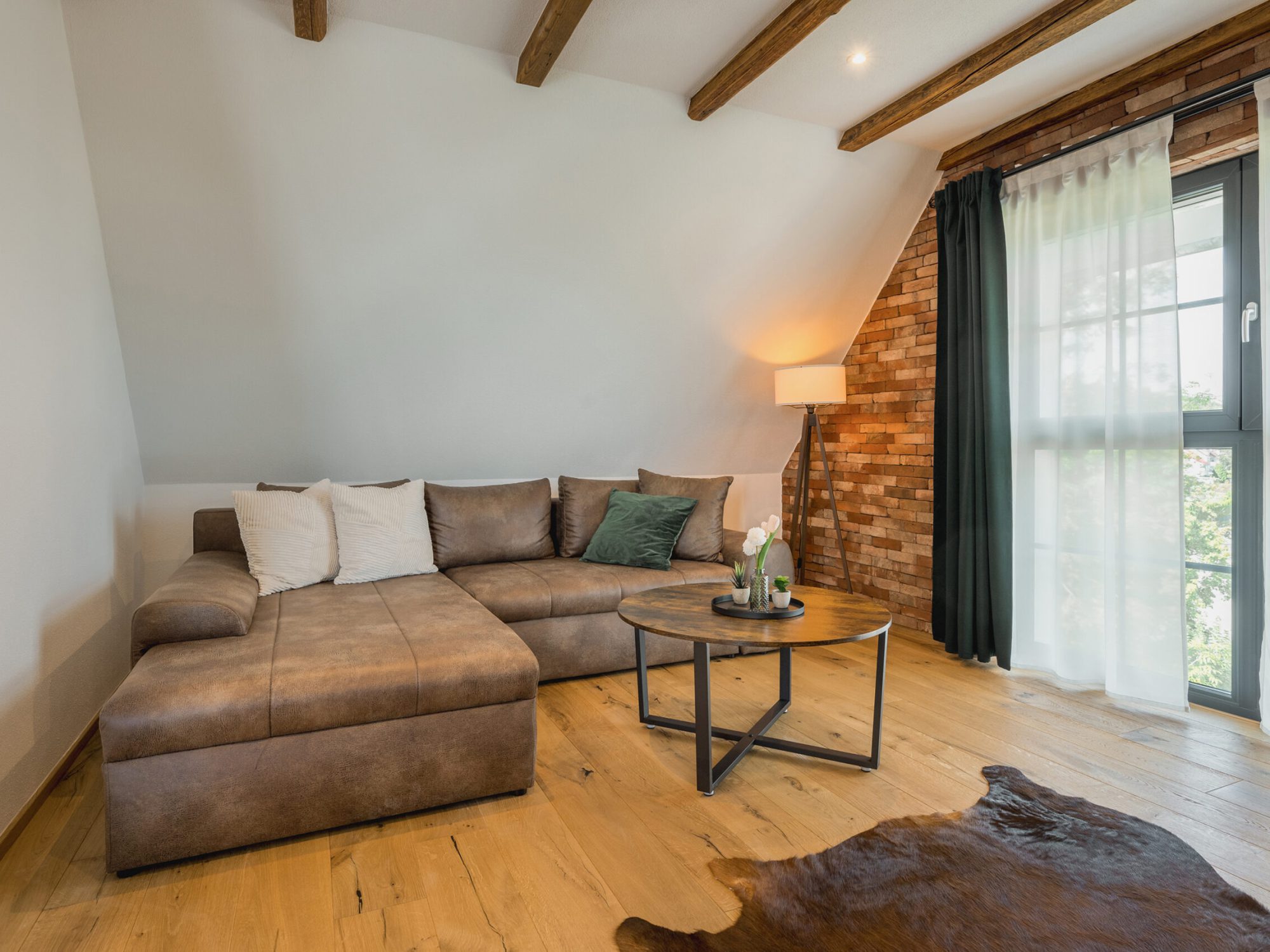 Braunes Sofa in Lederoptik in einem Raum mit Dachschräge und bodentiefen Fenster. Vor dem Sofa ein moderner Couchtisch in Holzoptik und schwarzen Stahlbeinen